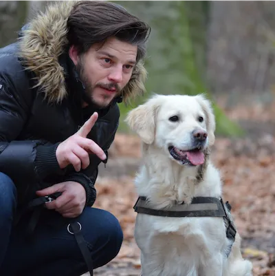 man with dark hair and facial hair crouched down and leaning in close to a white dog with a chest leash