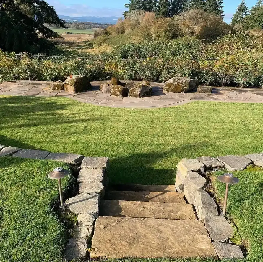 Natural flagstone patio with boulder fire ring, landscape lighting, stone steps, and Willamette Valley view by HD Landscape in Newberg