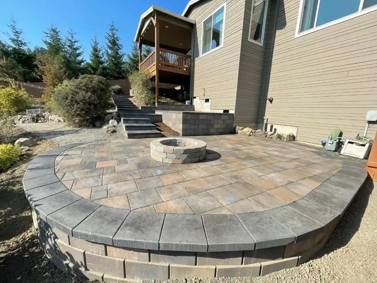 Raised circular paver patio with block fire pit and paver steps leading to wood deck by HD Landscape in Salem