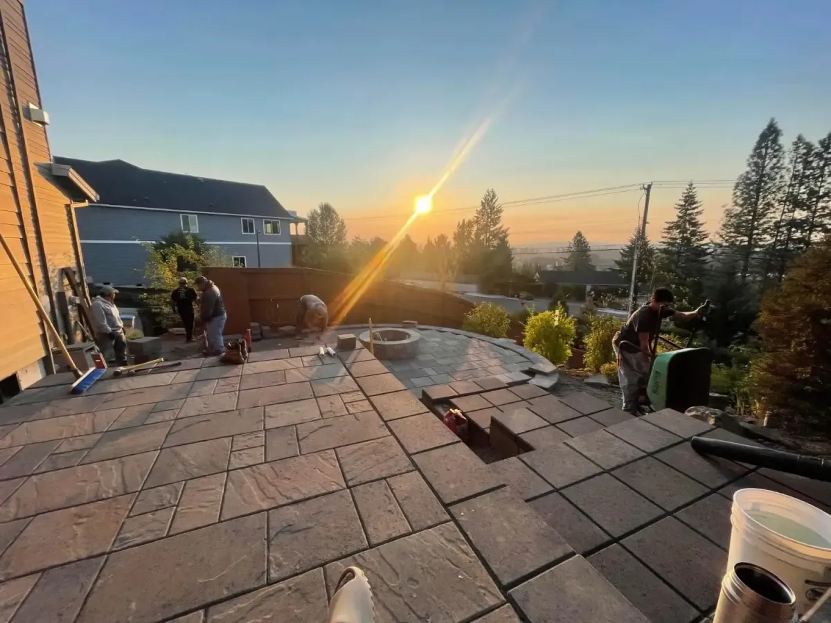 HD Landscape four-man crew finishing a circular paver patio and stone fire pit at sunset on a hillside property in West Linn