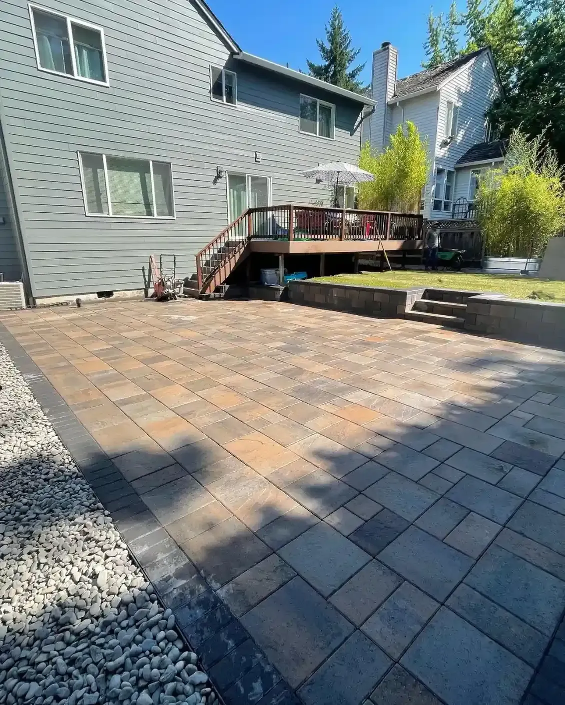Large paver patio with block retaining wall, steps, and river rock border beside wood deck by HD Landscape in Newberg
