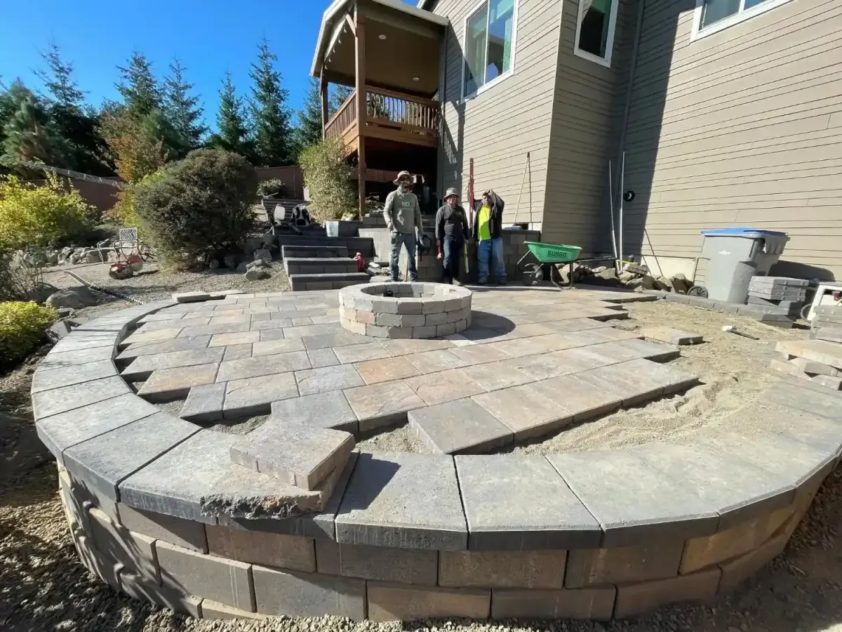 HD Landscape three-man crew beside a near-complete circular paver patio with fire pit ring and paver steps in West Linn