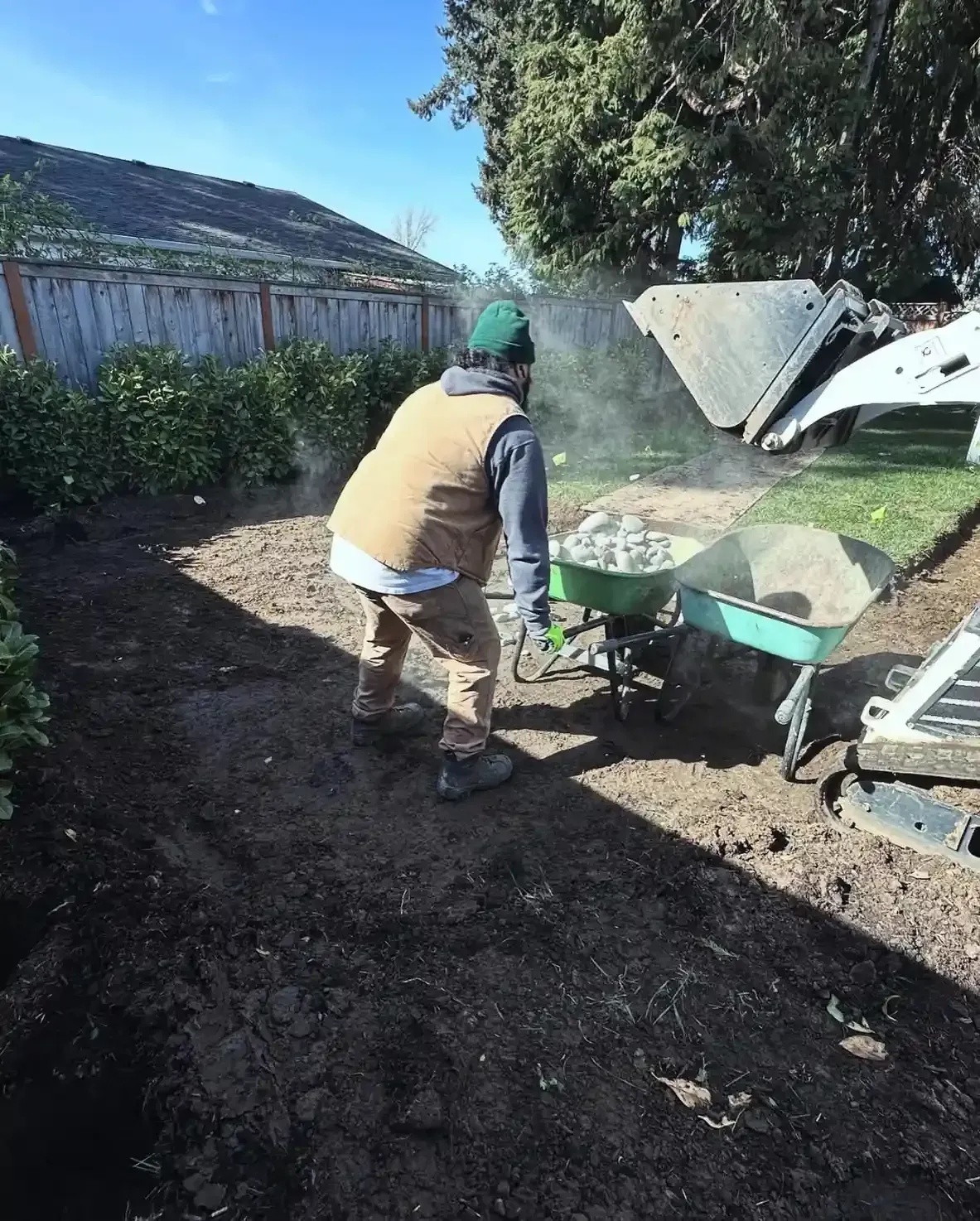 HD Landscape crew member loading river rock from a skid steer bucket into a wheelbarrow during a backyard installation in Portland