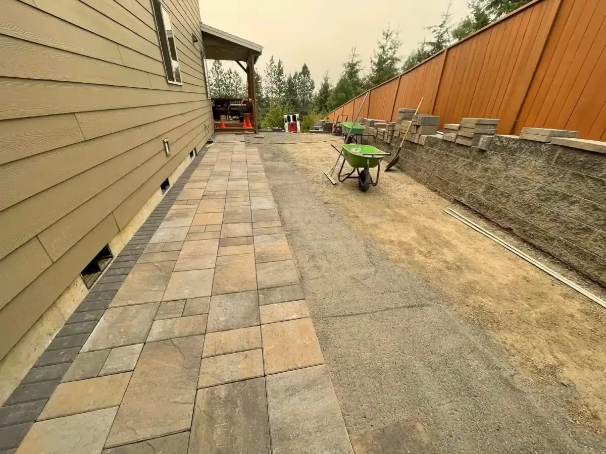 Paver patio installation in progress alongside a block retaining wall with staged materials by HD Landscape in Wilsonville