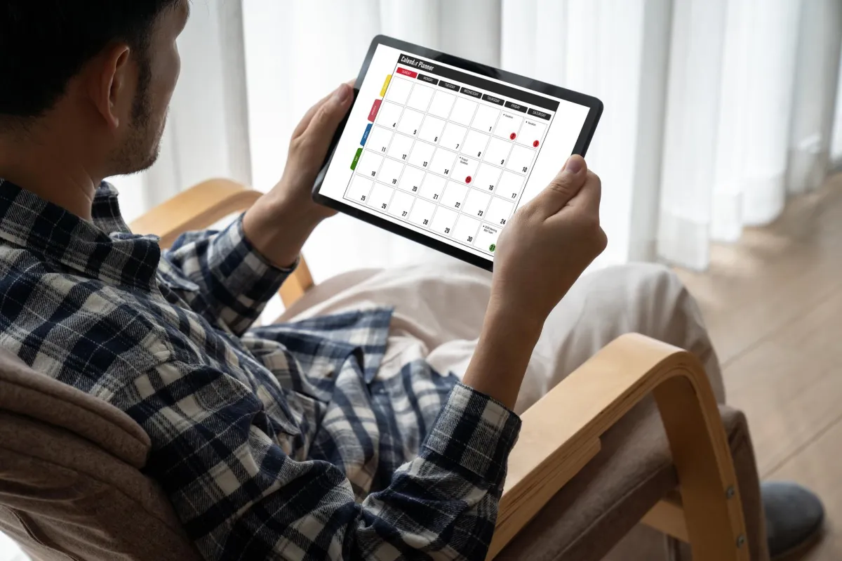 Man sitting on a chair holding a digital calendar tablet, planning future appointments or bookings.