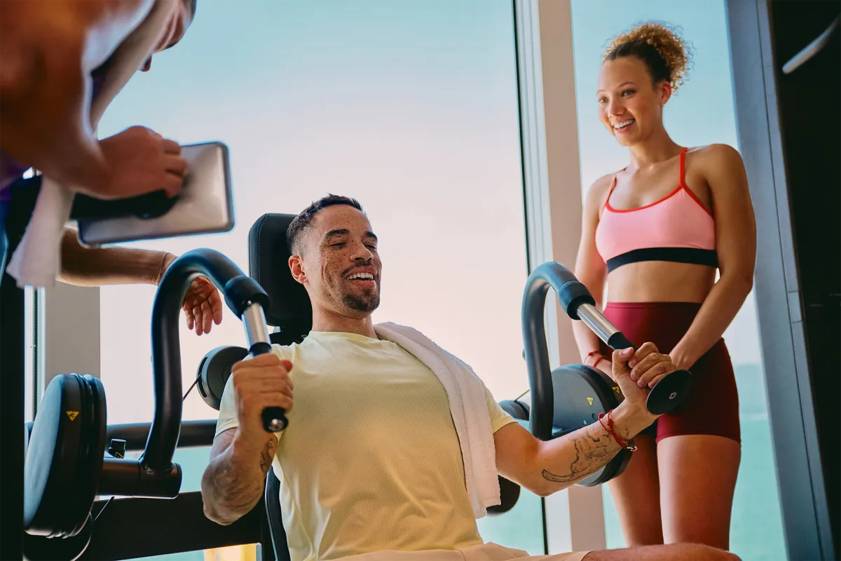 Man using strength training equipment in a modern gym with large ocean-view windows, accompanied by a smiling fitness instructor.