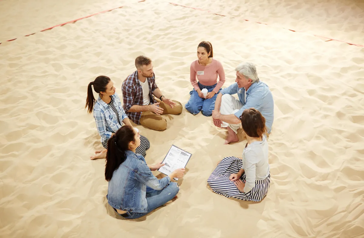 Group of adults sitting barefoot in a circle on a sandy beach, engaged in conversation during a curated excursion activity.