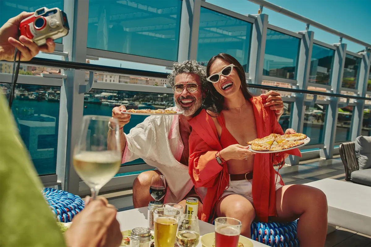 Smiling couple enjoying pizza and drinks on a sunny cruise deck, surrounded by friends and festive energy, capturing the relaxed and playful vibe of life at sea.