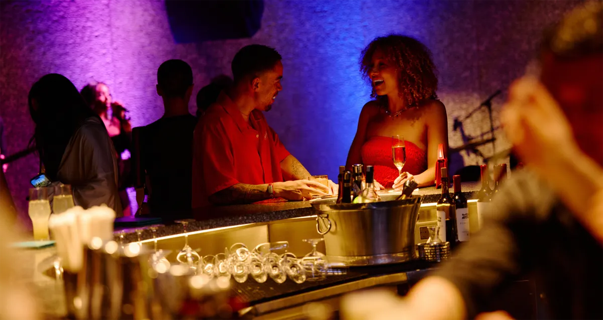 Guests socializing and enjoying drinks at a dimly lit bar with vibrant purple and blue party lighting, representing the exclusive VIP welcome celebration.