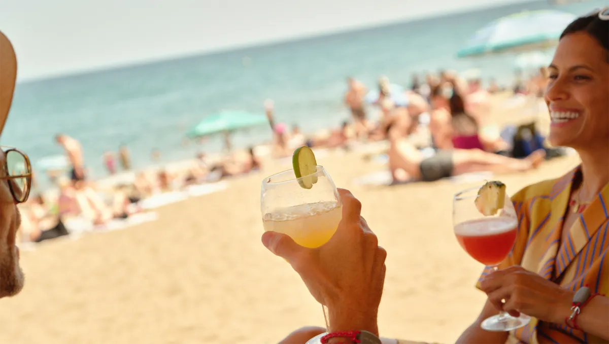 Two people smiling and toasting cocktails while lounging on a sunny beach with a crowd and umbrellas in the background.