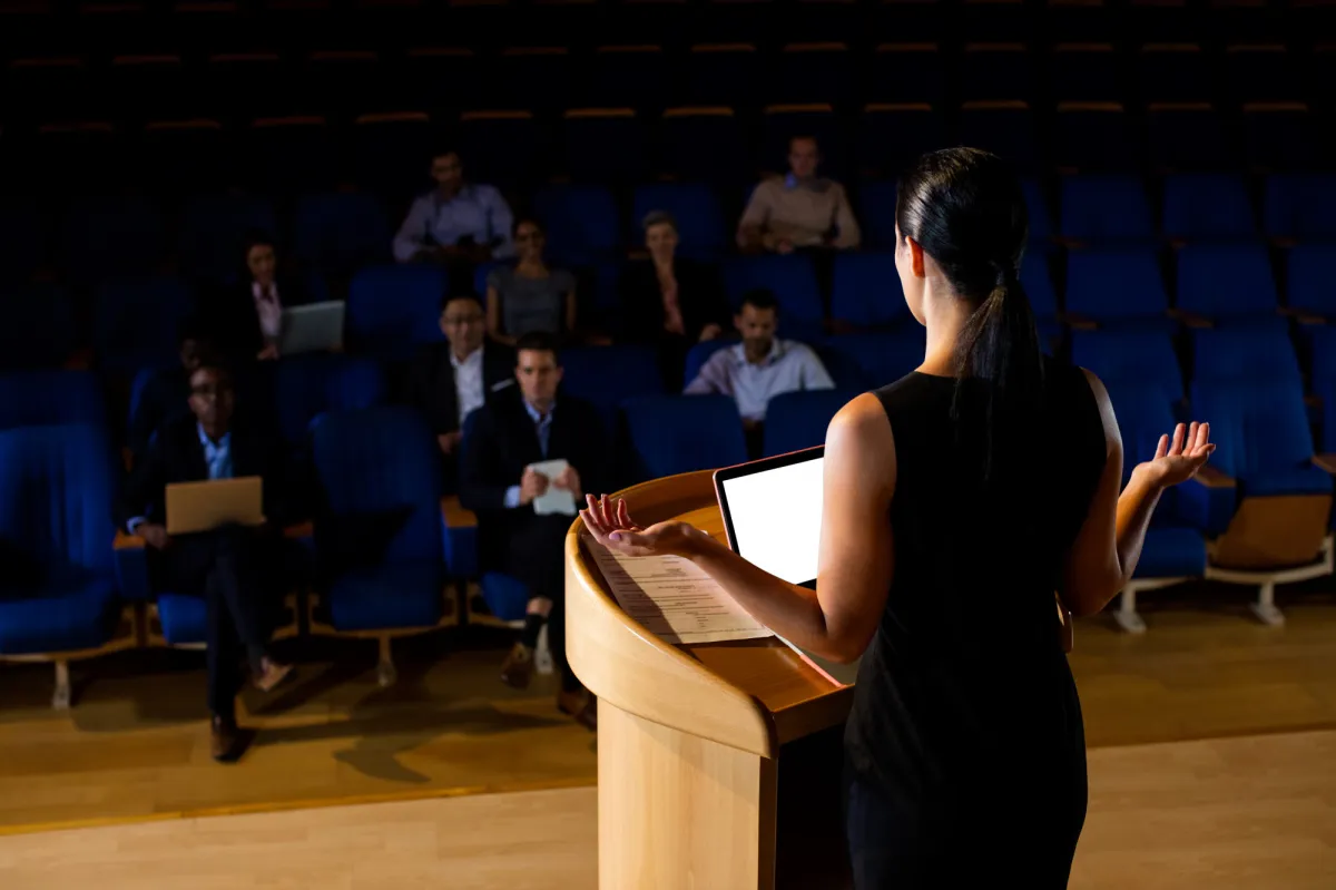 Female speaker delivering a talk at a podium to a small audience seated in a professional auditorium.