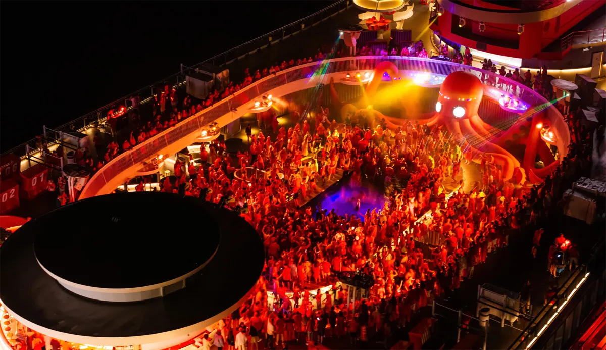 Aerial night view of a large crowd dancing at a colorfully lit deck party on the Virgin Voyages ship.