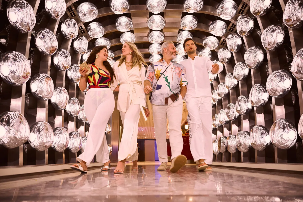 Group of stylish cruise guests walking through a futuristic, mirrored hallway filled with glowing light fixtures, showcasing the luxurious atmosphere aboard the ship.
