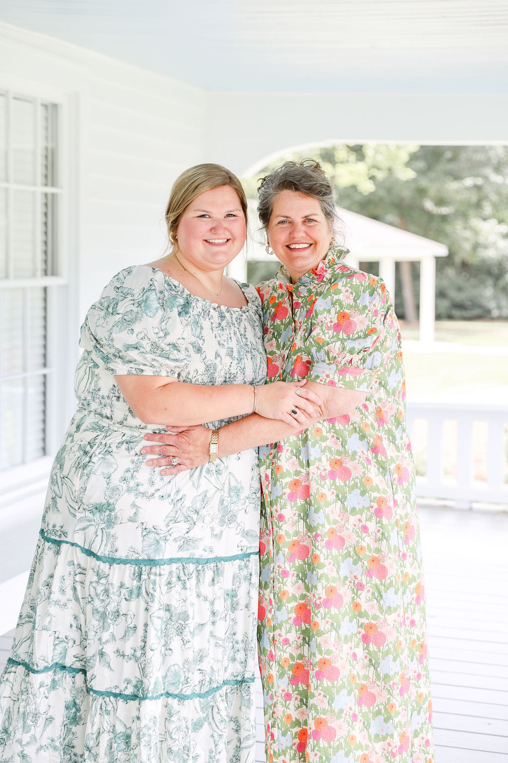 Two women standing outdoors, holding hands, smiling, with a field and overcast sky in the background.