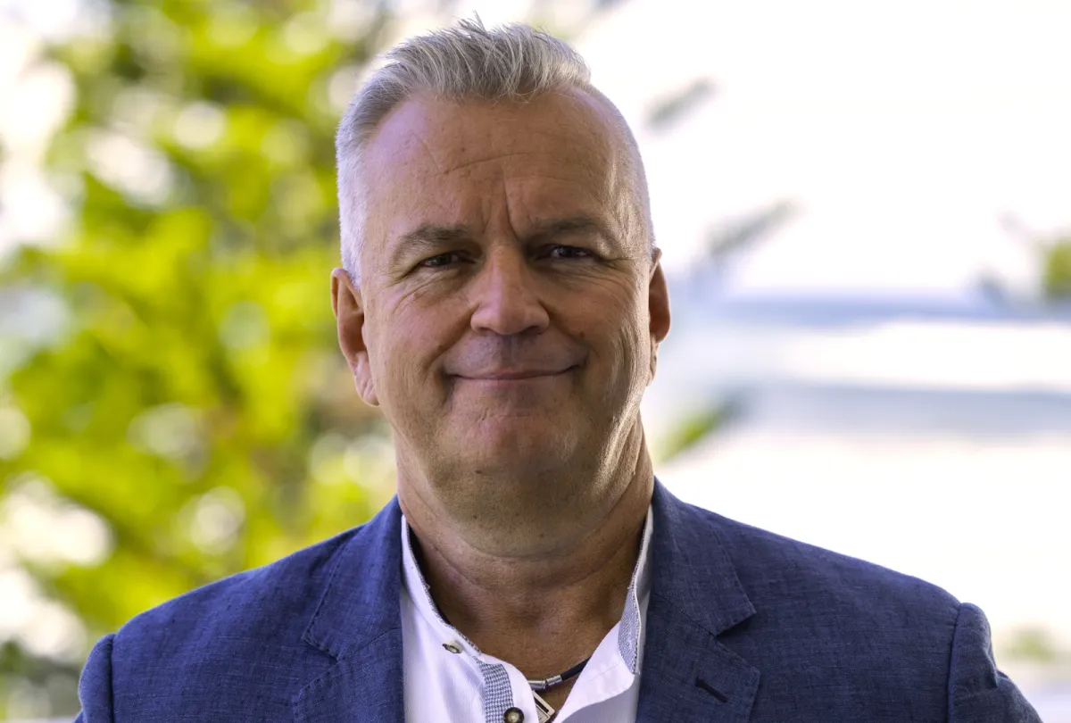 Mark Robinson wearing a navy blazer and white shirt, standing outdoors with greenery in the background and smiling at the camera.