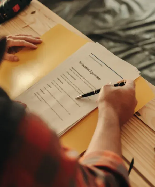 A person in a plaid shirt reviews a 'Renovation Agreement' document with a pen in hand on a wooden table.