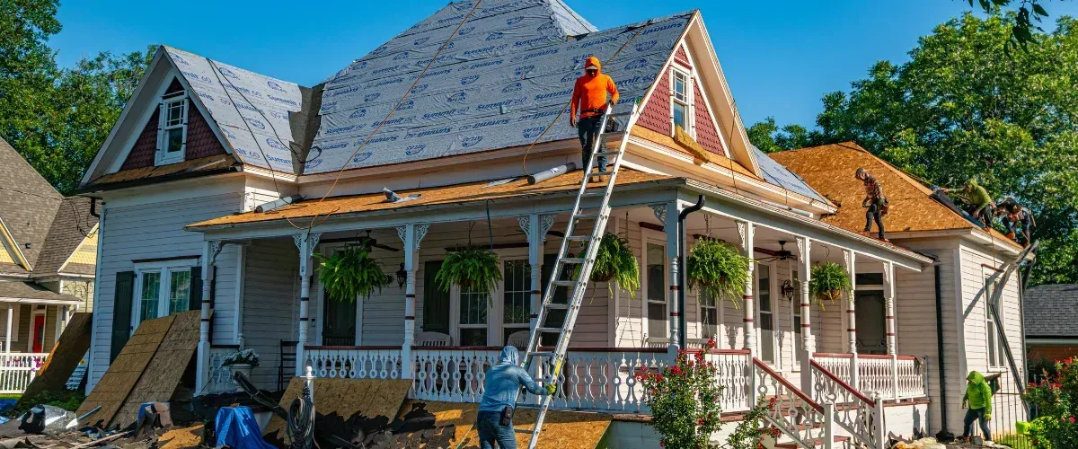 Several roofers work on a large historic two-story house with a partially exposed roof and blue sky overhead.