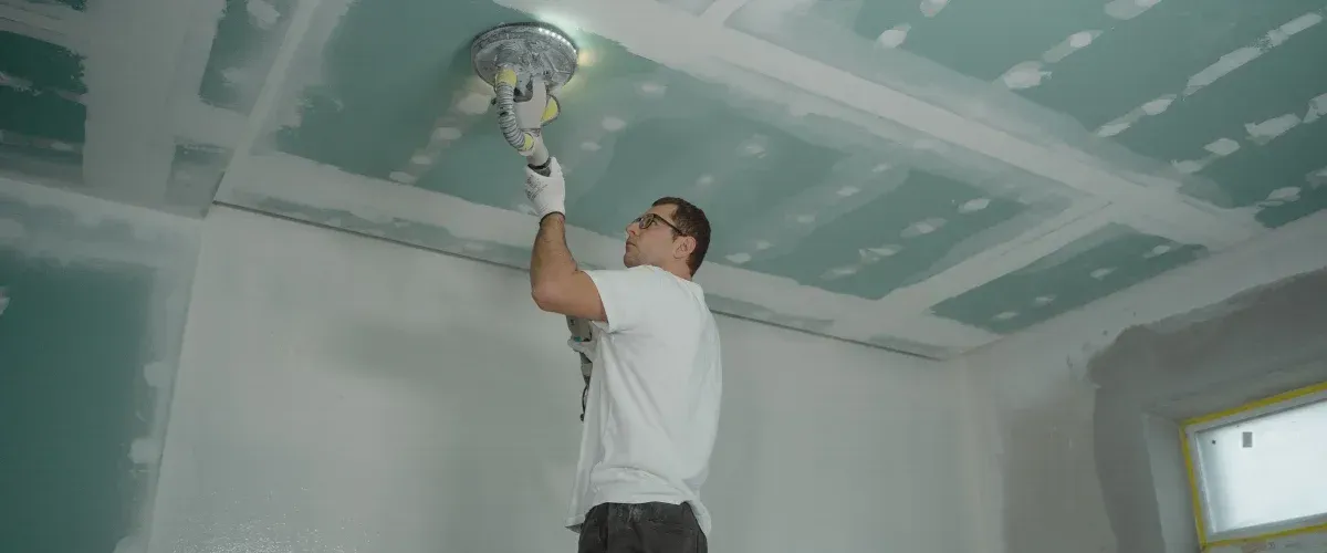 A man wearing glasses and a white t-shirt uses an orbital sander on a drywall ceiling in a room under construction.