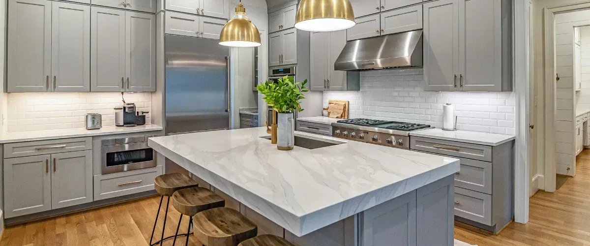 A wide angle view of an elegant kitchen with gray cabinetry, a large marble island, and stainless steel appliances.