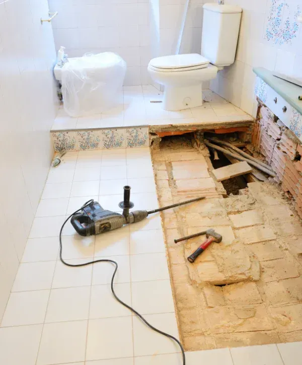 A bathroom undergoing renovation with a jackhammer on the tiled floor and a section of the floor removed.