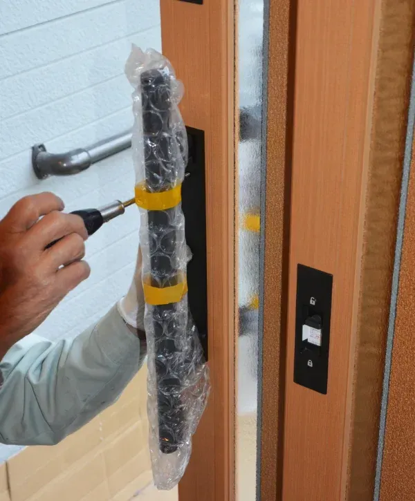A person installs a new door handle, still wrapped in bubble wrap, onto a light wooden door with a screwdriver.