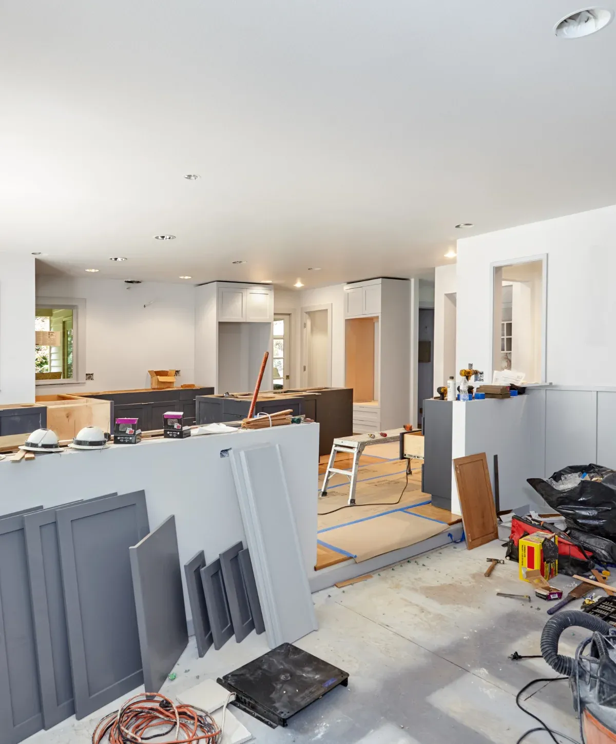 A wide shot of a home kitchen and living area under construction with gray cabinets, white upper cabinets, and tools scattered.