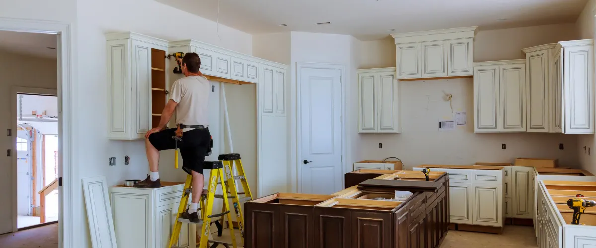 A man on a ladder installs white upper kitchen cabinets with a drill, while lower cabinets are set in an unfinished room.