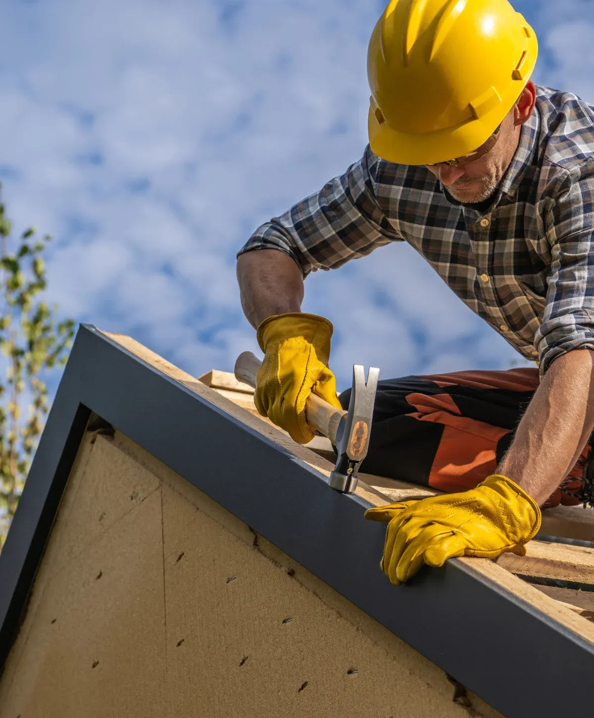 A construction worker in a plaid shirt and tan cap crouches on a roof installing dark gray tiles, with scaffolding visible left.