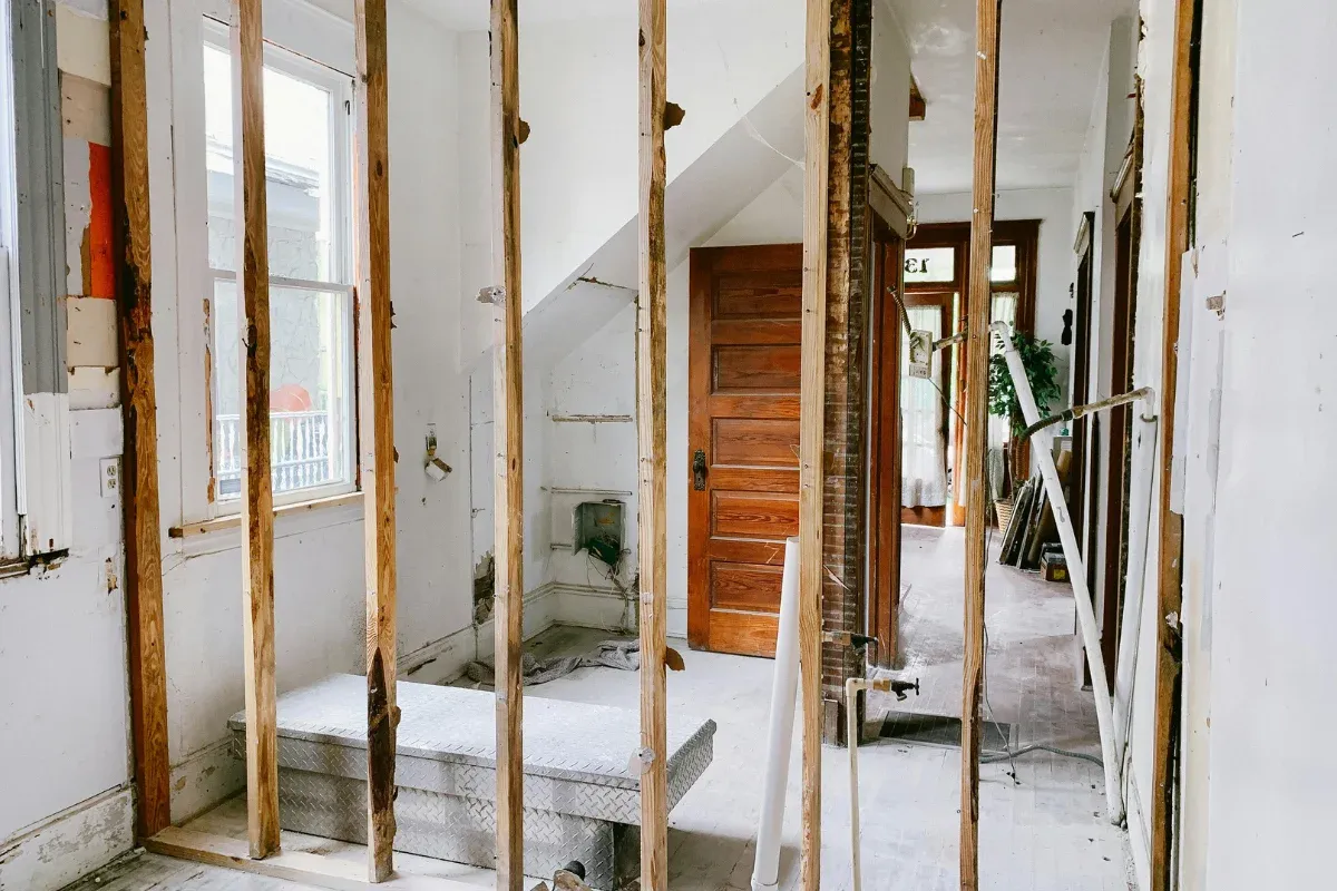 An interior view of a house undergoing renovation, with exposed wooden wall studs, a window, and a hallway beyond.