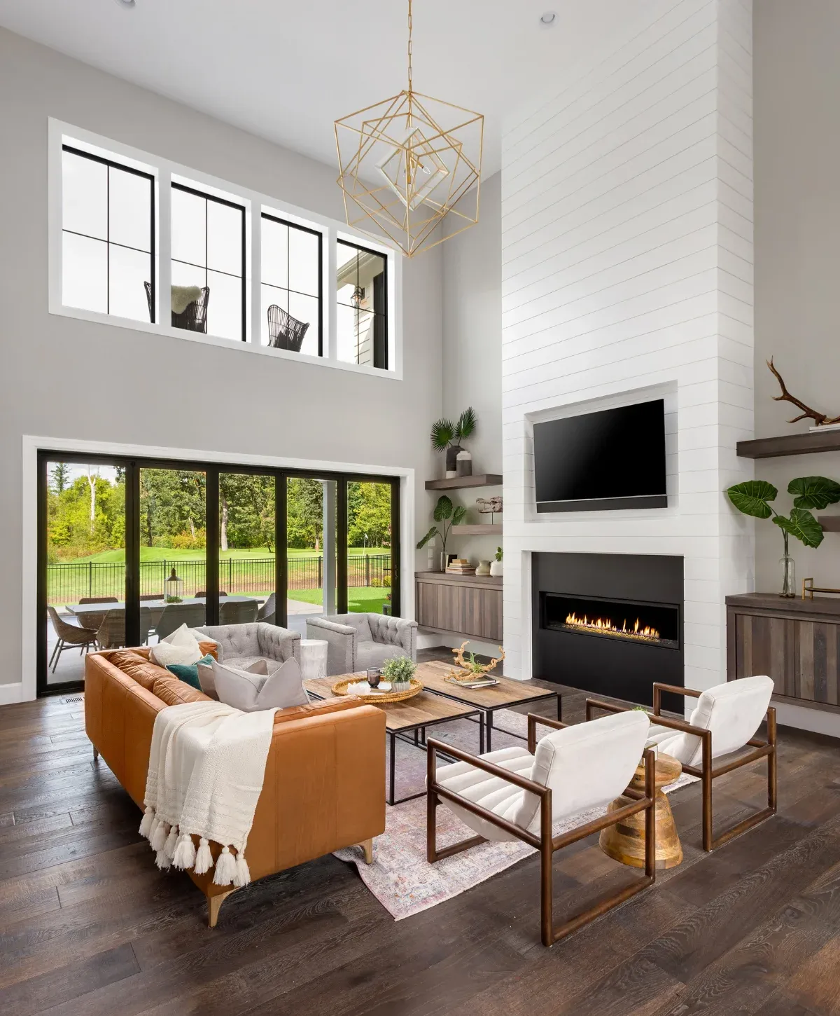 A modern, open-concept living room with a tall fireplace, large windows overlooking a green yard, and a brown leather sofa.