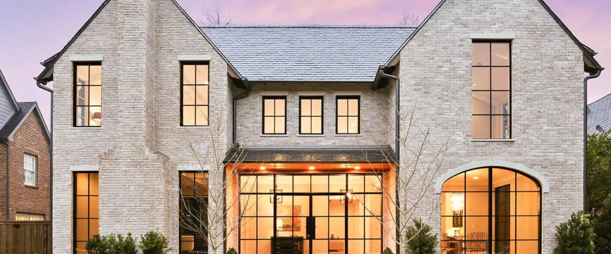 A modern, light-colored brick house with large black-framed windows and gabled roofs under a purple sky at dusk.