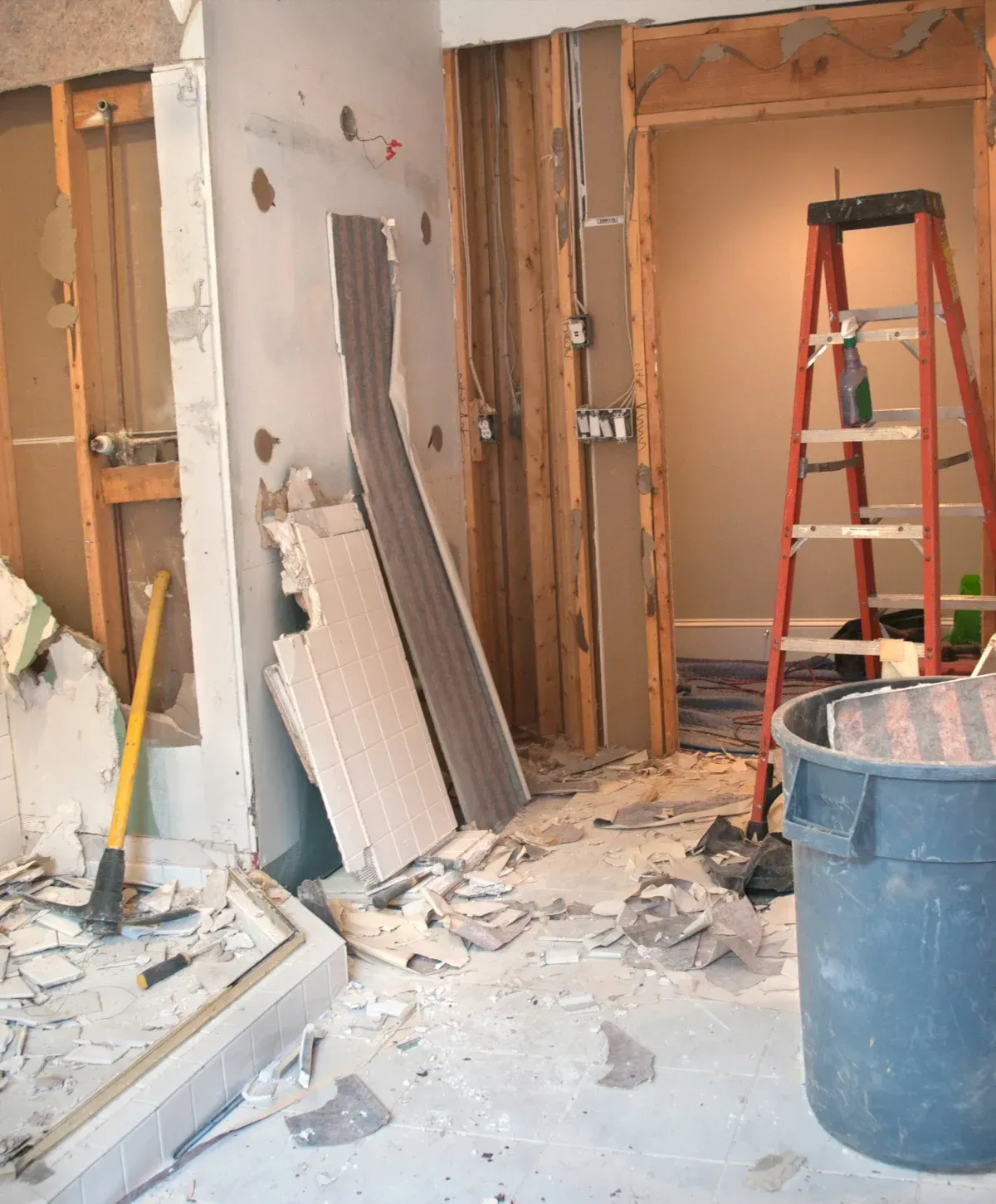 A room undergoing demolition with exposed wall studs, scattered tile and drywall debris, a yellow sledgehammer, a red ladder, and a grey trash can.