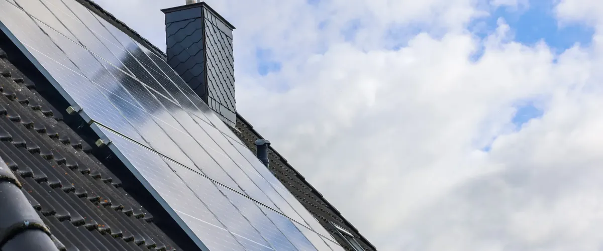 Solar panels installed on a dark tiled roof next to a chimney, viewed against a cloudy sky.