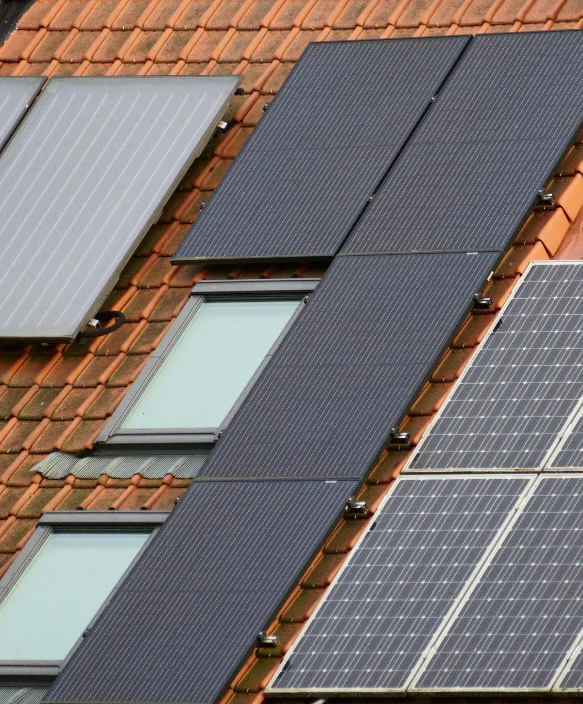 An aerial close-up of dark solar panels, a thermal collector, and two skylight windows on a terracotta tiled roof.