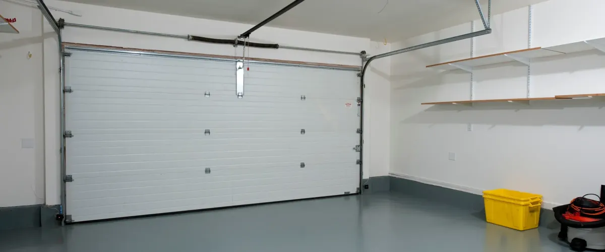 A clean, well-lit garage interior with a closed white sectional door, gray epoxy floor, and empty wall shelves.