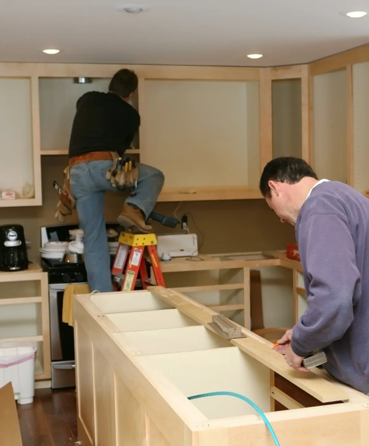Two men are actively installing light wood kitchen cabinets and a central island in a home renovation project.