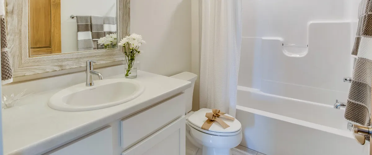 A bright, modern bathroom featuring a white vanity with sink, a framed mirror, a toilet with a burlap bow, and a white shower-tub combo.