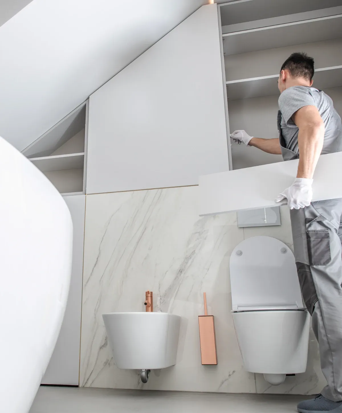 A person installs a white cabinet in a modern attic bathroom with a freestanding tub, bidet, and toilet.