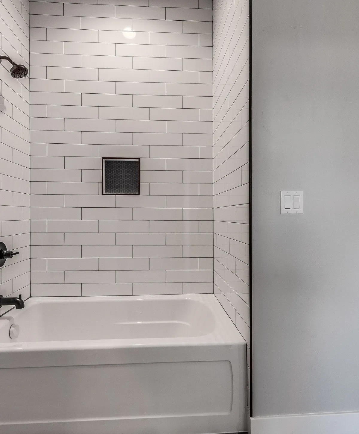 A modern bathroom features a white bathtub against white subway tiles with a dark recessed shower niche and bronze fixtures.