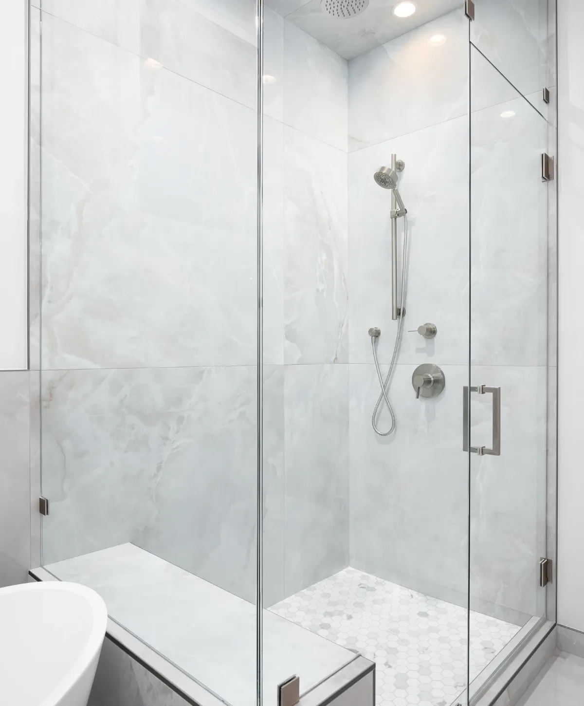 A luxurious modern walk-in shower with clear glass doors, light marble-look tiles, a built-in bench, and hexagonal floor.