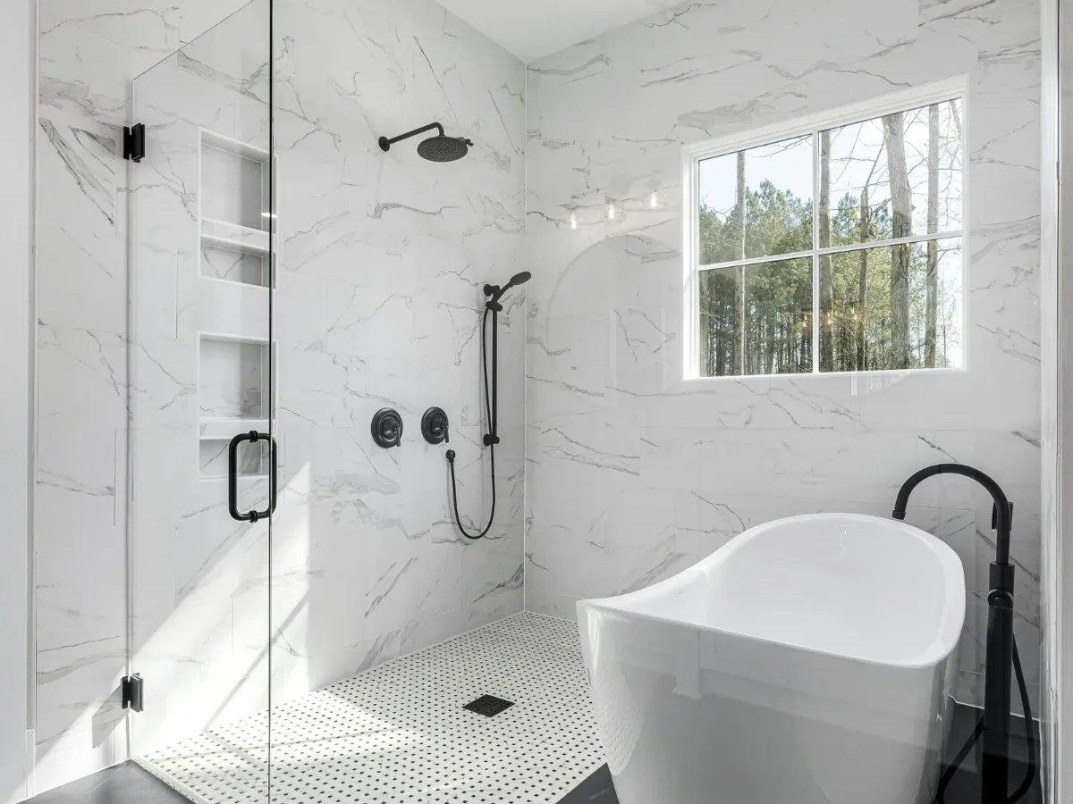A luxurious modern bathroom featuring white marble-look tiles, a glass shower with black fixtures, and a freestanding bathtub by a window.