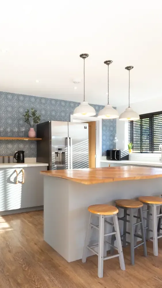 Contemporary kitchen with blue geometric feature wall, white island with oak breakfast bar, wooden bar stools, and white pendant lights by Blueprint Kitchens