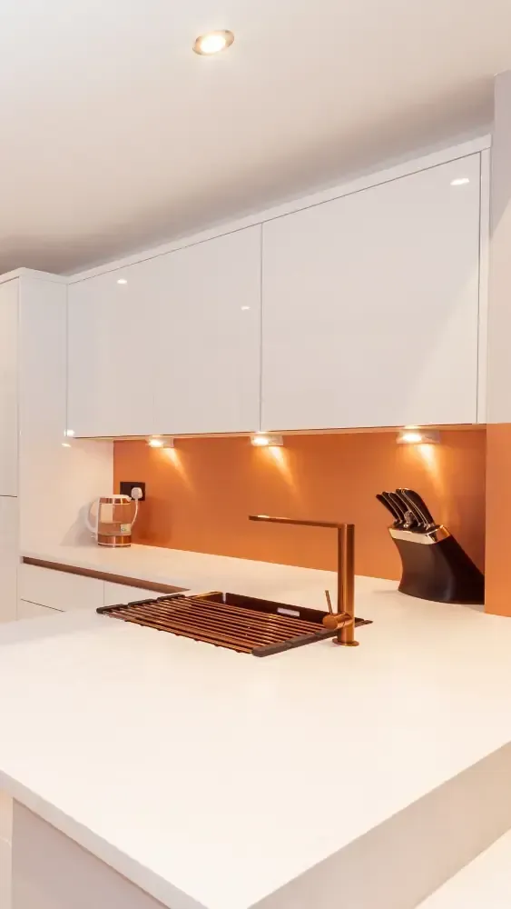 Modern white kitchen with bright orange backsplash, copper tap and drying rack, handleless cabinets, and LED under-cabinet lighting by Blueprint Kitchens