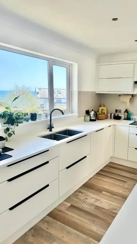 White handleless kitchen with black accent strips, corner window overlooking sea view, black tap, and wood flooring by Blueprint Kitchens