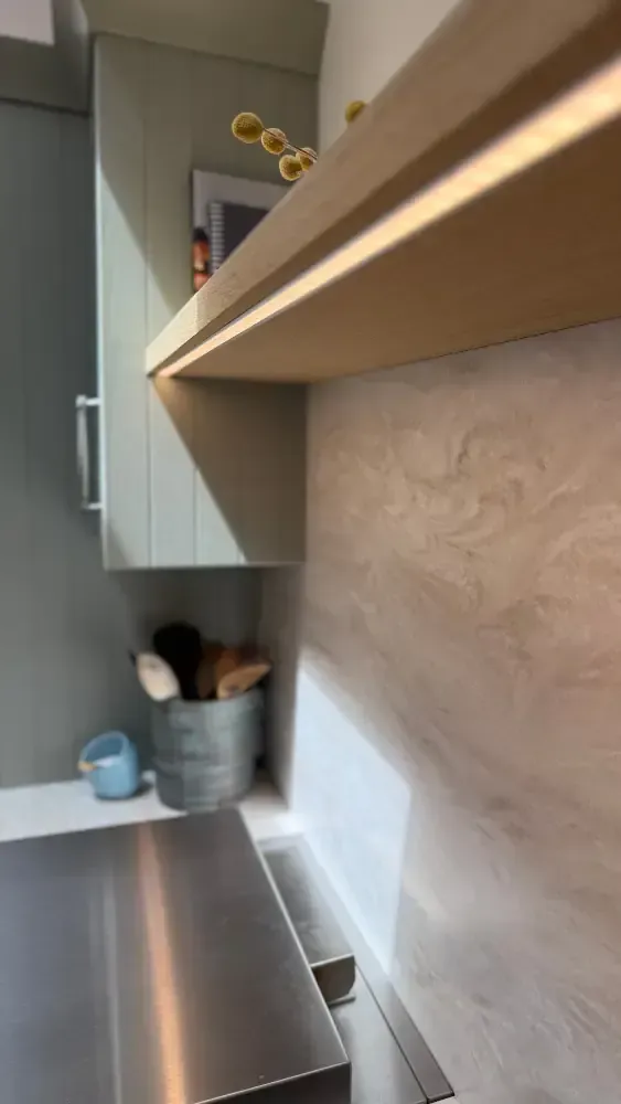 Oak floating shelf with integrated LED lighting, textured backsplash, sage green cabinets, and kitchen accessories by Blueprint Kitchens