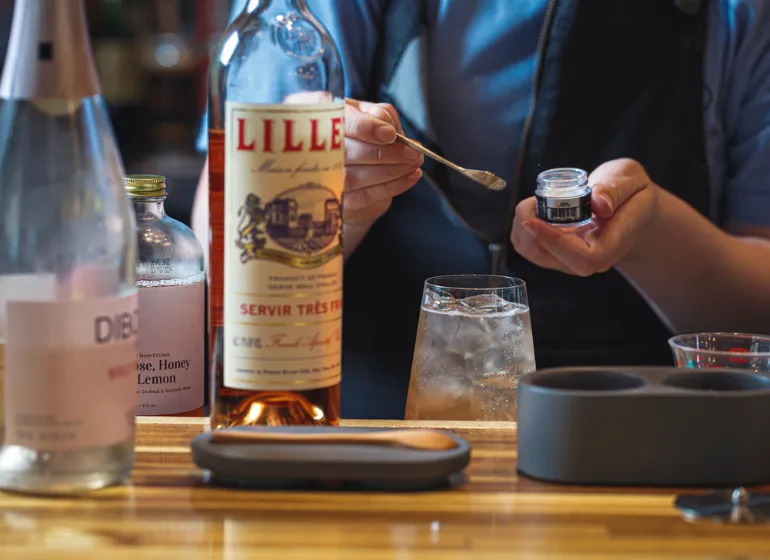 Bartender crafting a specialty cocktail at Stroll Cafe, adding edible beverage glitter alongside premium ingredients like Lillet and botanical mixers.