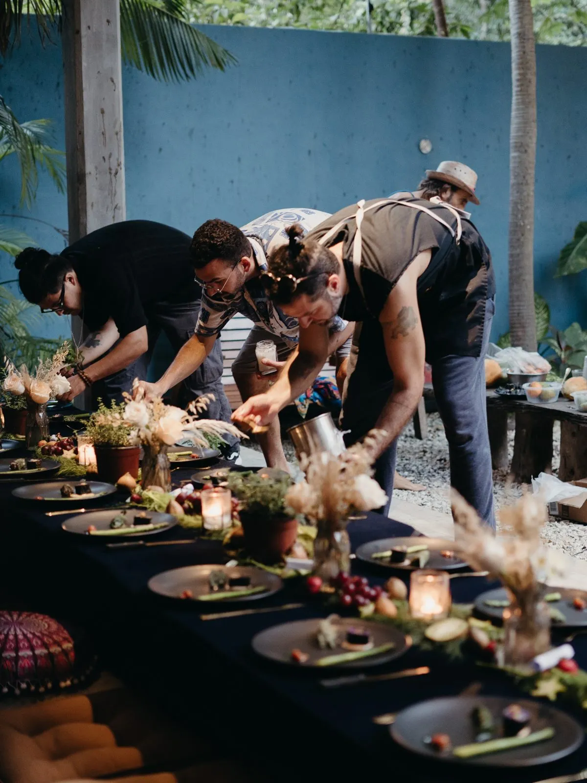 A long outdoor dinner table being set with plates of fresh food