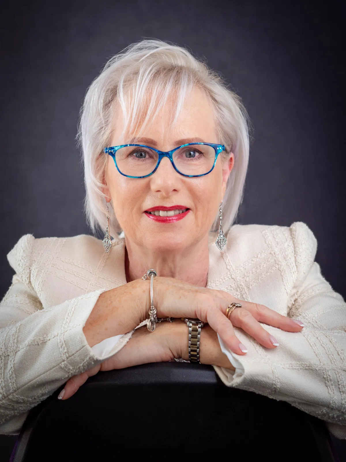 Jackie is a passionate mastermind leaning on folded arms that are resting on the back of a chair She has bright blue glasses on and a cream blazer. facilitator and coach for small business owners who want to grow confidently and sustainably. She is wearing a cream blazer and bright blue glasses. Her arms are folded and leaning on the back of a chair.