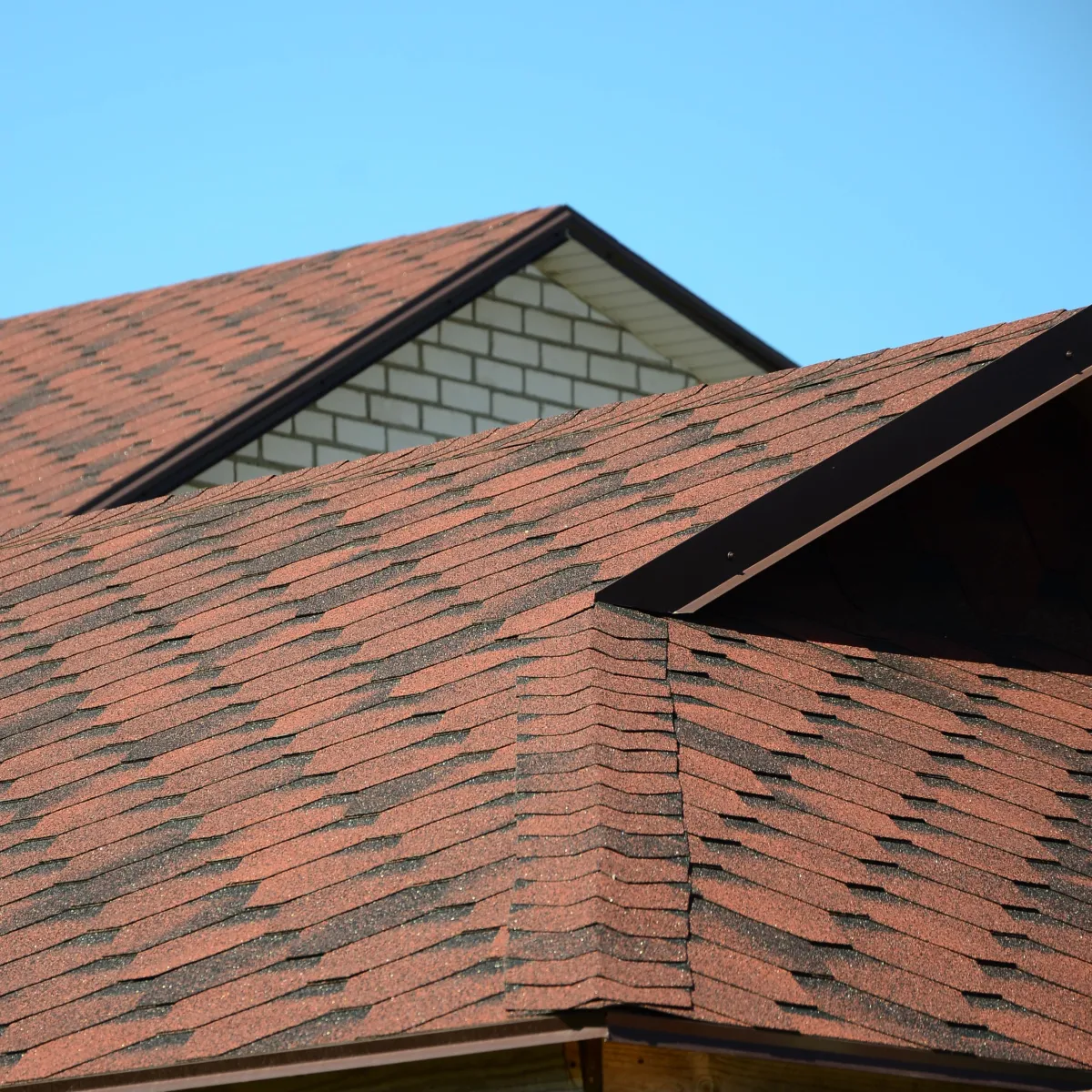 Red asphalt shingle roof on a house with brick walls under clear blue sky.