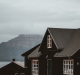 Dark wooden house with white windows and a mountain backdrop under cloudy sky.